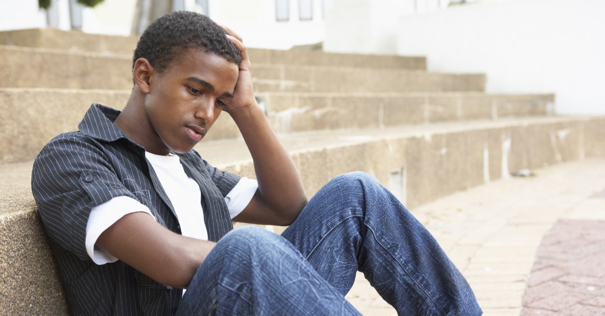 A teenager sitting on steps, looking down with a hand on his head, experiencing anxiety and depression.