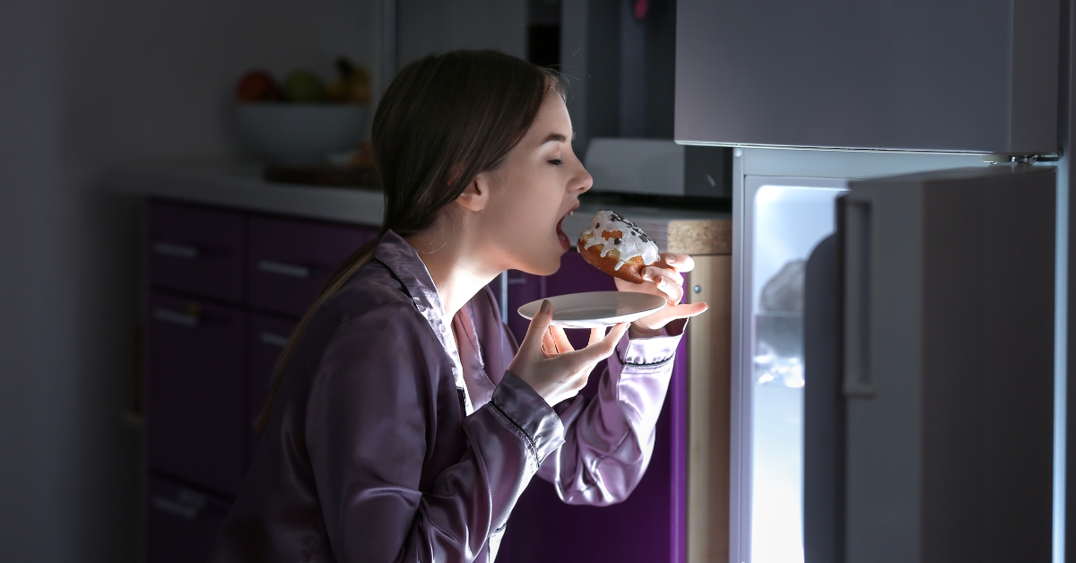 A woman eating a pastry in front of an open fridge, highlighting tips to stop binge eating at night.