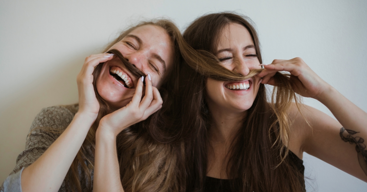 Two women laughing and playfully using their hair as mustaches, illustrating Common Reasons for Fear of Being Happy.