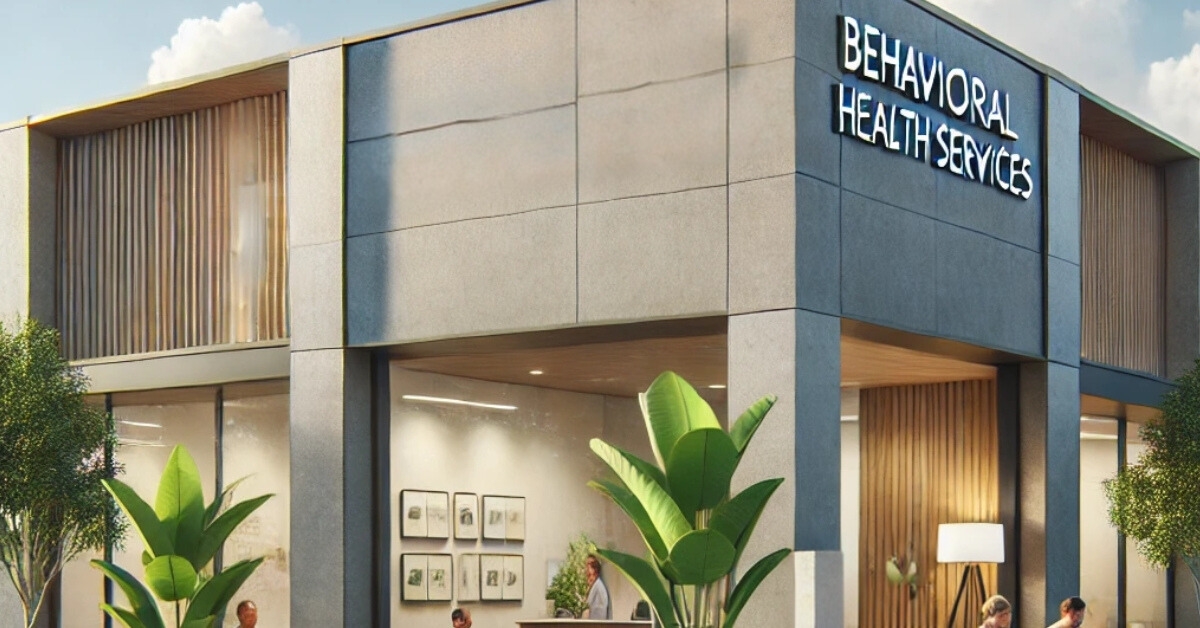 A modern building with large windows displays a sign for "Behavioral Health Services." The welcoming entrance has plants and people inside.