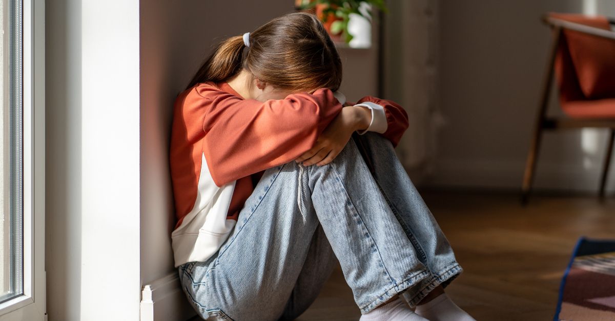 A young woman sits on the floor with her head buried in her arms, looking distressed. She wears a red sweater and jeans.