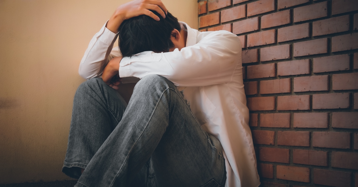 A teenager sitting against a wall, head down and holding their head, illustrating stress in teens.