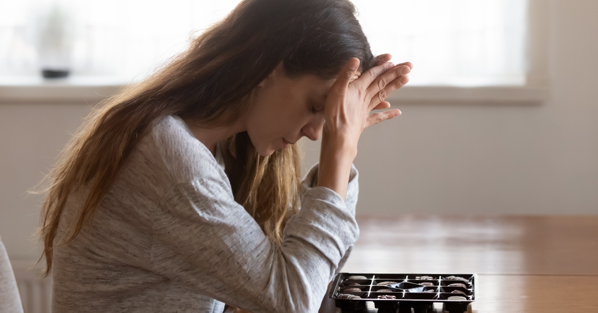 A woman sits at a table with her head in her hands, looking at a box of chocolates, illustrating ARFID for adults.