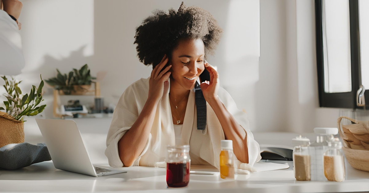 A woman with curly hair smiles while talking on the phone, surrounded by wellness items including a laptop, jars, and a plant.