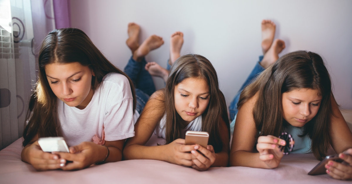 Three teenage girls lying on a bed, engrossed in their phones, illustrating the impact of social media on a teen’s sense of self naturally.