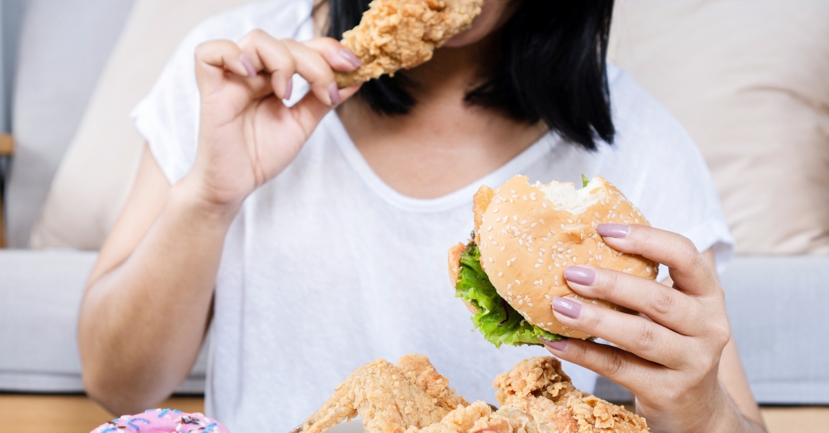 A woman eating a burger and fried chicken with more food in front of her, illustrating Binge Eating.