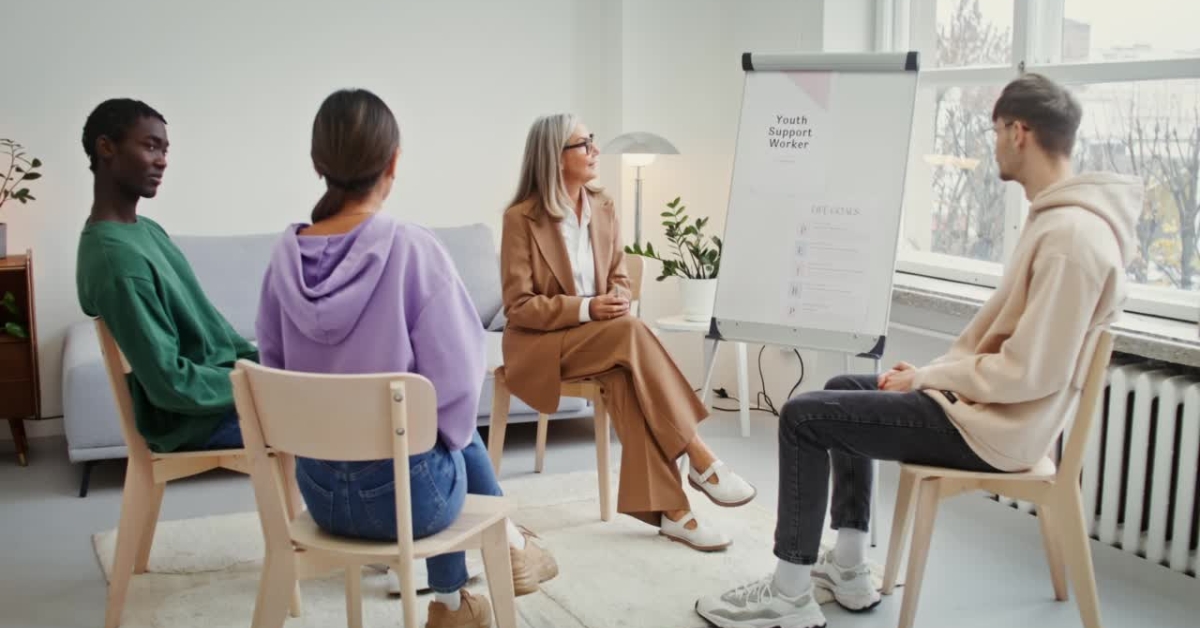 A group of young adults in a session with a therapist, discussing mood therapy in a bright room.