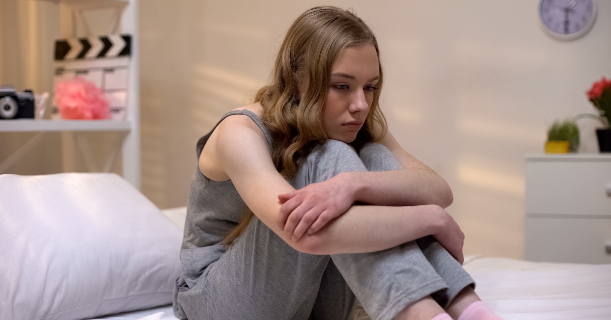 Teen girl sitting on a bed, hugging her knees, looking sad, depicting separation anxiety in teens.